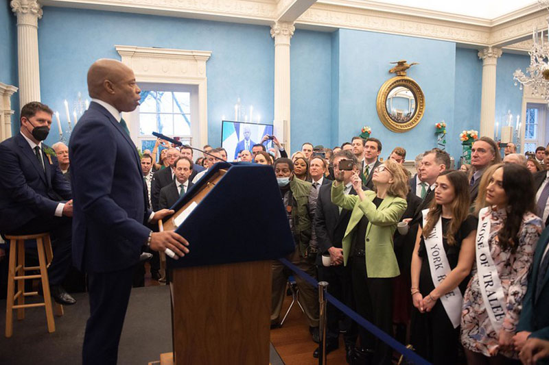 Mayor Eric Adams stands onstage behind his podium addressing guests. Minister Eamon Ryan, Ireland's Minister for Environment, Climate, and Communications & Minister for Transport sits behind him on a stool.