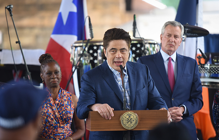 Benicio del Toro speaking from the podium on stage with the Mayor and First Lady standing behind him