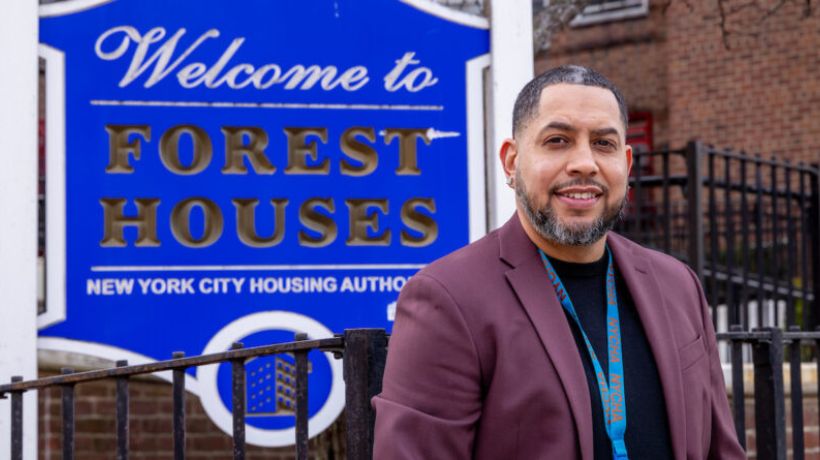 Portrait of NYCHA Superintendent Edwin Pagan smiling at a work cubicle, wearing a blue NYCHA lanyard over his Bermuda graphic t-shirt.

                                           