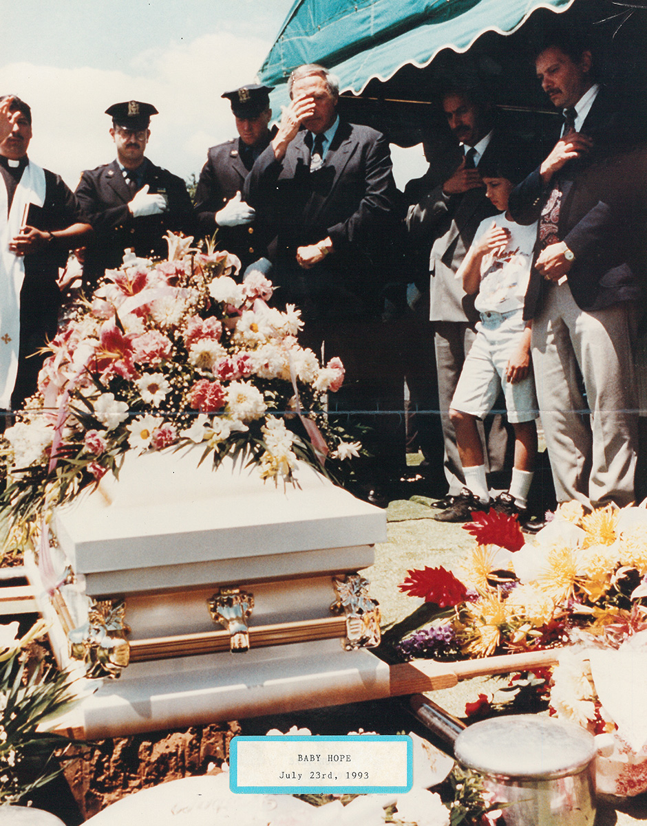 Small, white coffin before being lowered, cops stadnding at attention as a priest jesters a blessing