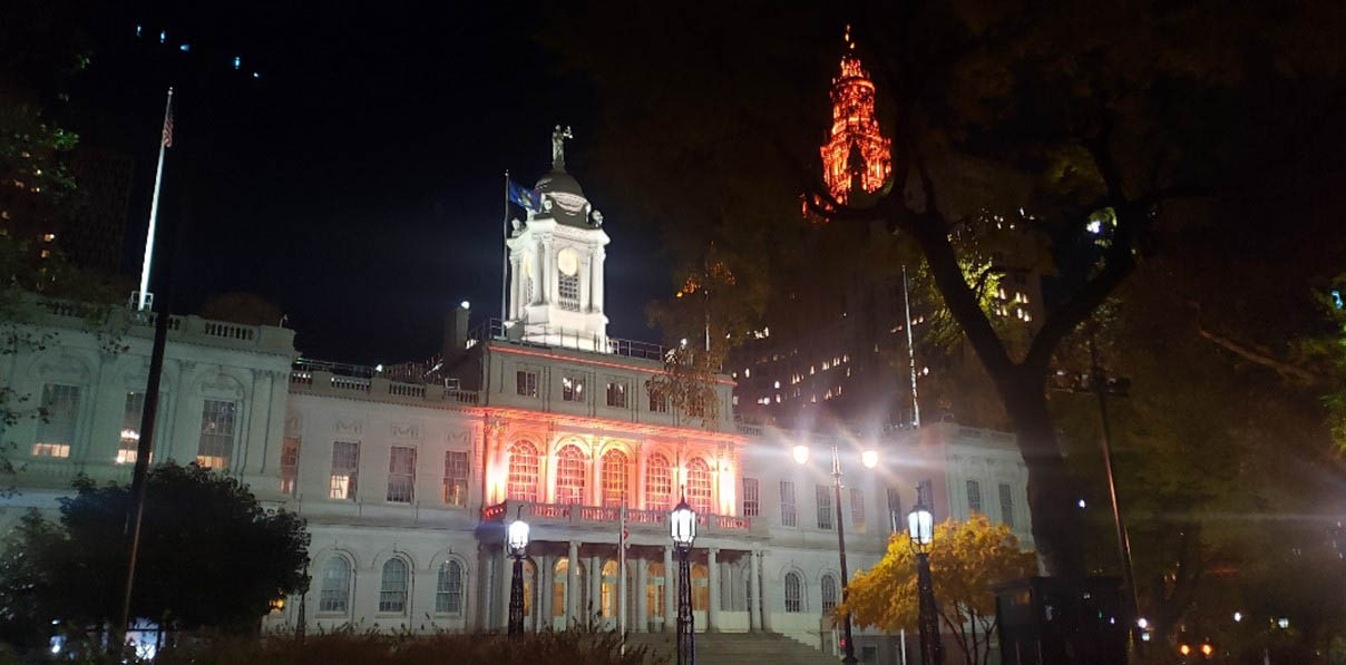 Outside view of NY City Hall Building illuminated in orange lights.
