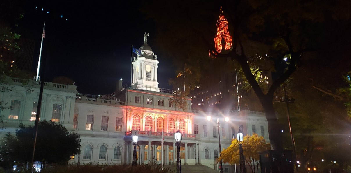 Outside view of NY City Hall Building illuminated in orange lights.
