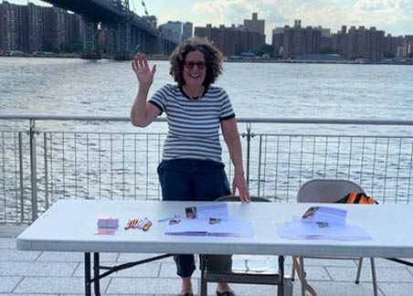 One woman in a striped shirt standing behind a white table with pamphlets at a riverfront promenade. 