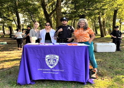 Three people and one NYC Police Officer standing behind a purple tablecloth covered table with pamphlets in an outdoor park.