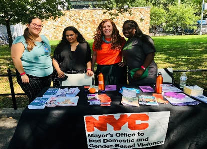 Four women standing behind a black tablecloth covered table with pamphlets in an outdoor park.
