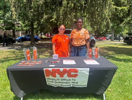Two men women in orange t-shirts standing behind a table filled with handouts/giveaways in outdoor park, Staten Island NY.