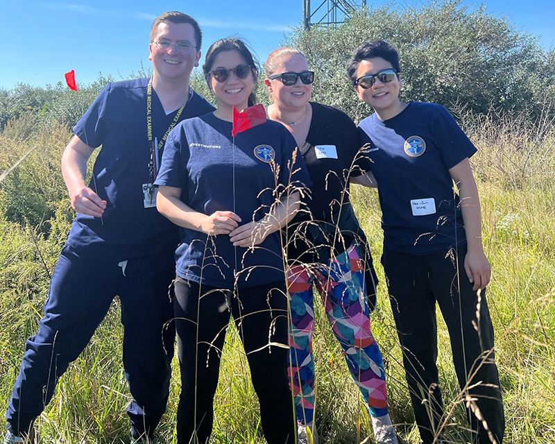 Four forensic pathology fellows from OCME, who are medical examiners in training, pose in the field on a sunny day during an outdoor training exercise. 