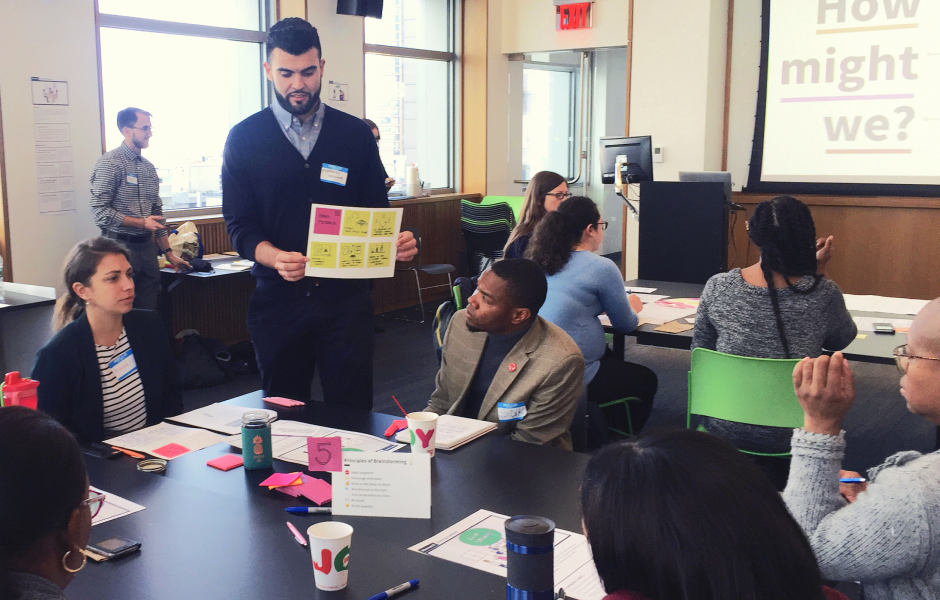 A group of people sitting around a table and a man explaining the notes he made on a sign with post-it notes.
