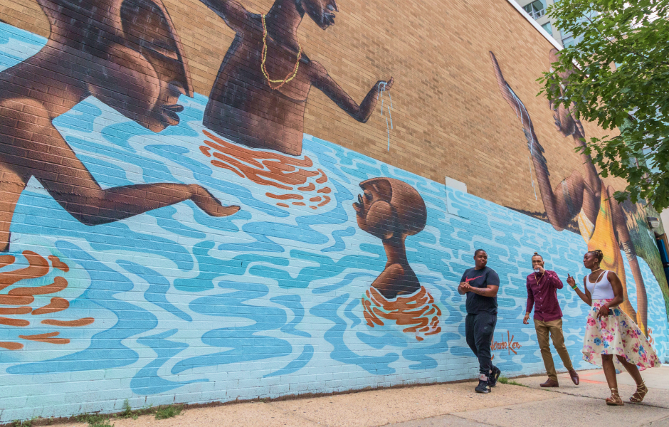 3 people walking down the street with a large mural on the side of a building.