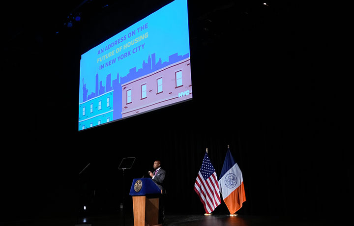 NYC Mayor Adams Speaking with a screen with illustration of skyline 
                                           