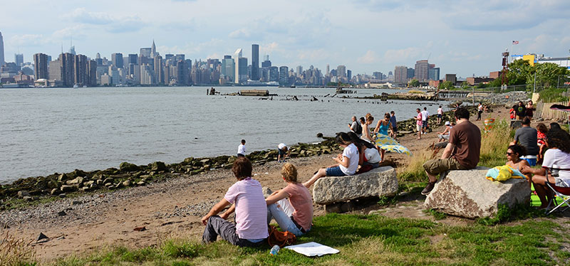 Groups of people sit alongside the shore with a view of the skyline in the background.