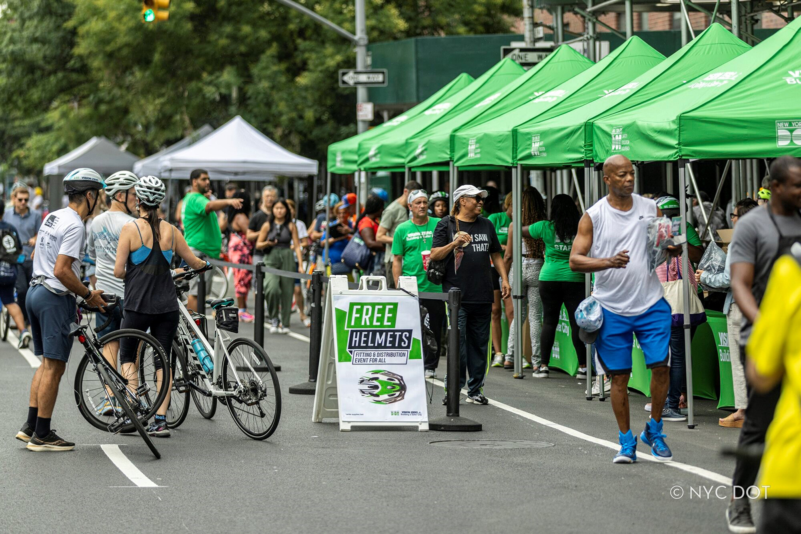 A busy city street fair features multiple green tents where people gather for a community event