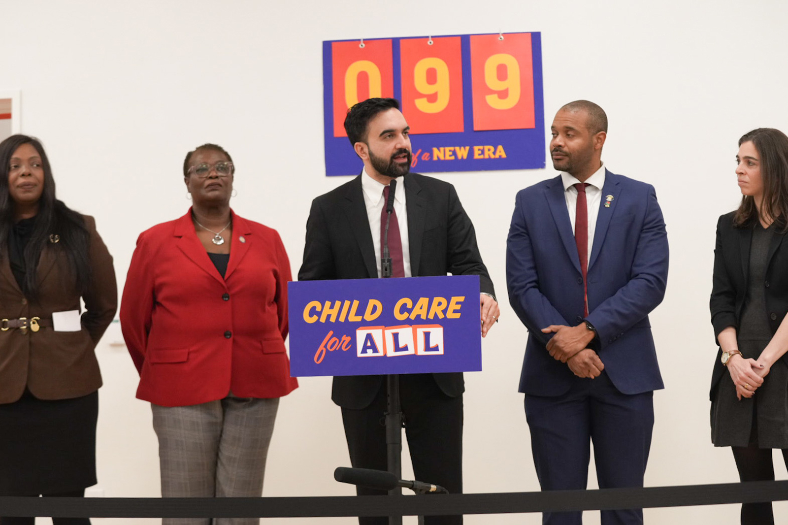 Mayor Mamdani stands in front of a podium with a sign reading 'Child Care for All.' 