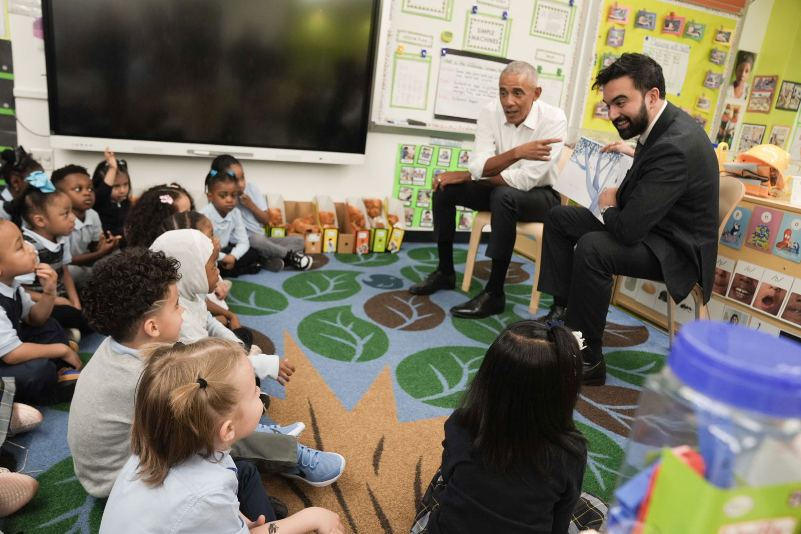 Former President Obama and Mayor Mamdani are presenting a story from a children's book, while a group of kids sit in the front and litening attentively.