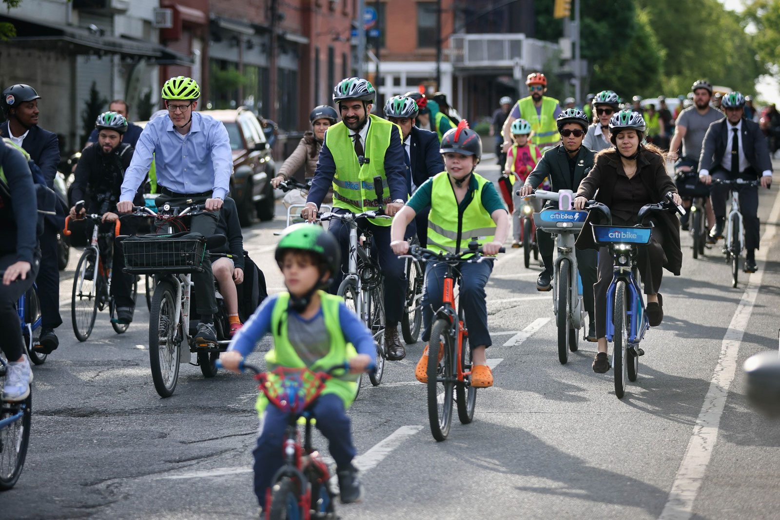 Mayor Mamdani in the center, wear green short vest and bike helmet, is among a group of people riding bicycle on the street