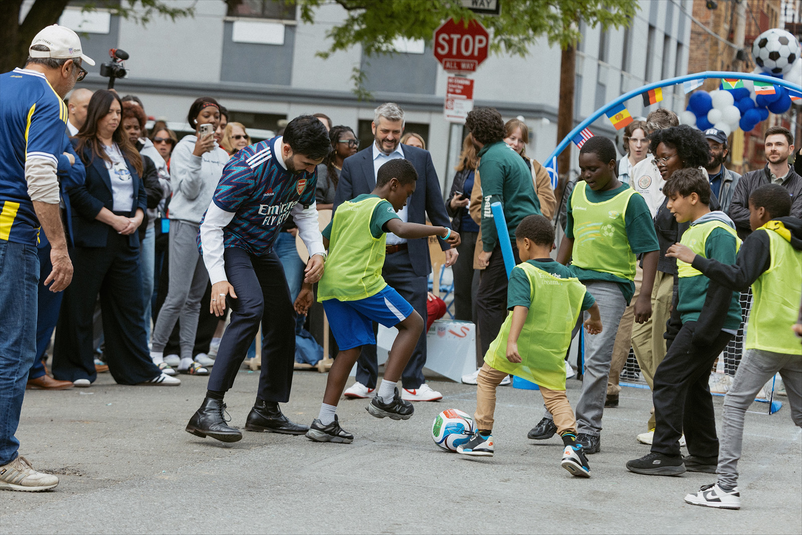 Mayor Mamdani and children wearing green pinnies playing soccer on the street