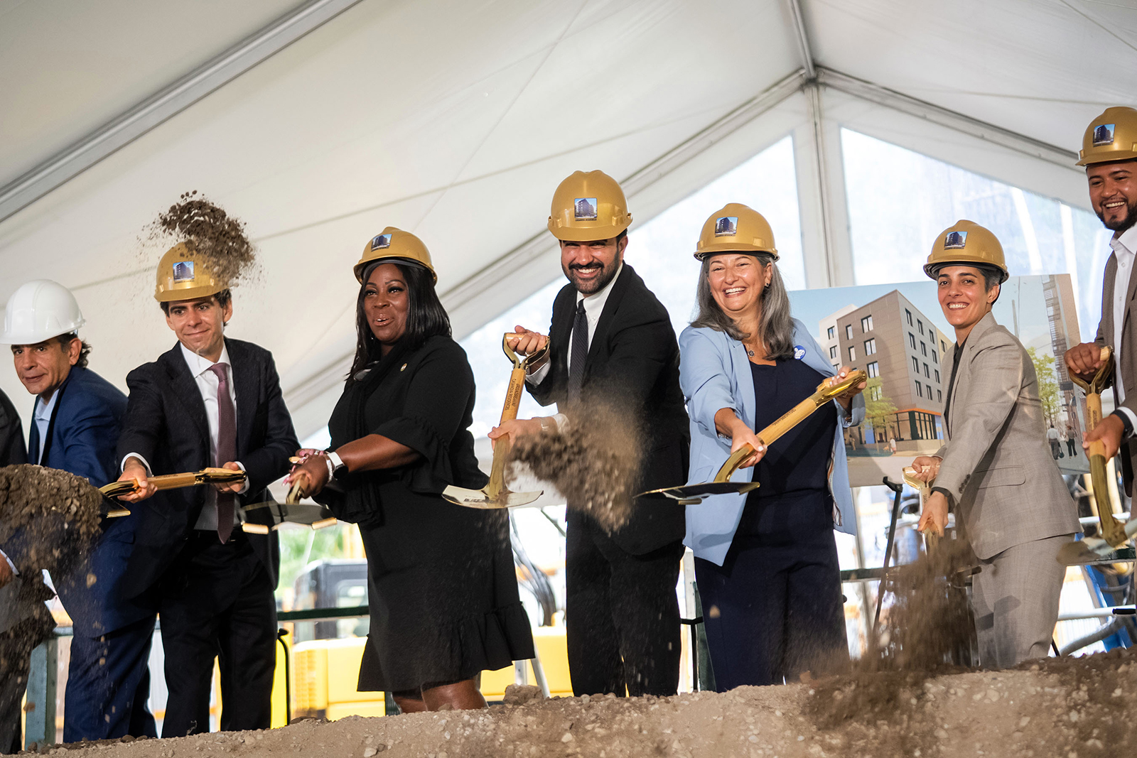 Mayor Mamdani and a diverse group of people in business attire and safety helmets participate in a groundbreaking ceremony. They stand under a large white tent, each holding a shovel and tossing dirt into the air. Construction equipment and a building rendering are visible in the background, suggesting a new development project. The scene conveys collaboration, progress, and community investment.