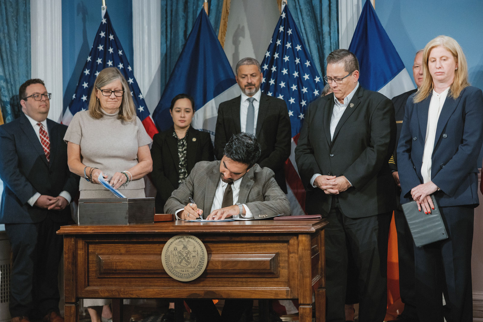 Mayor Mamdani, in gray suit, seated, head tilted, with his right hand holds a pen, signing a document