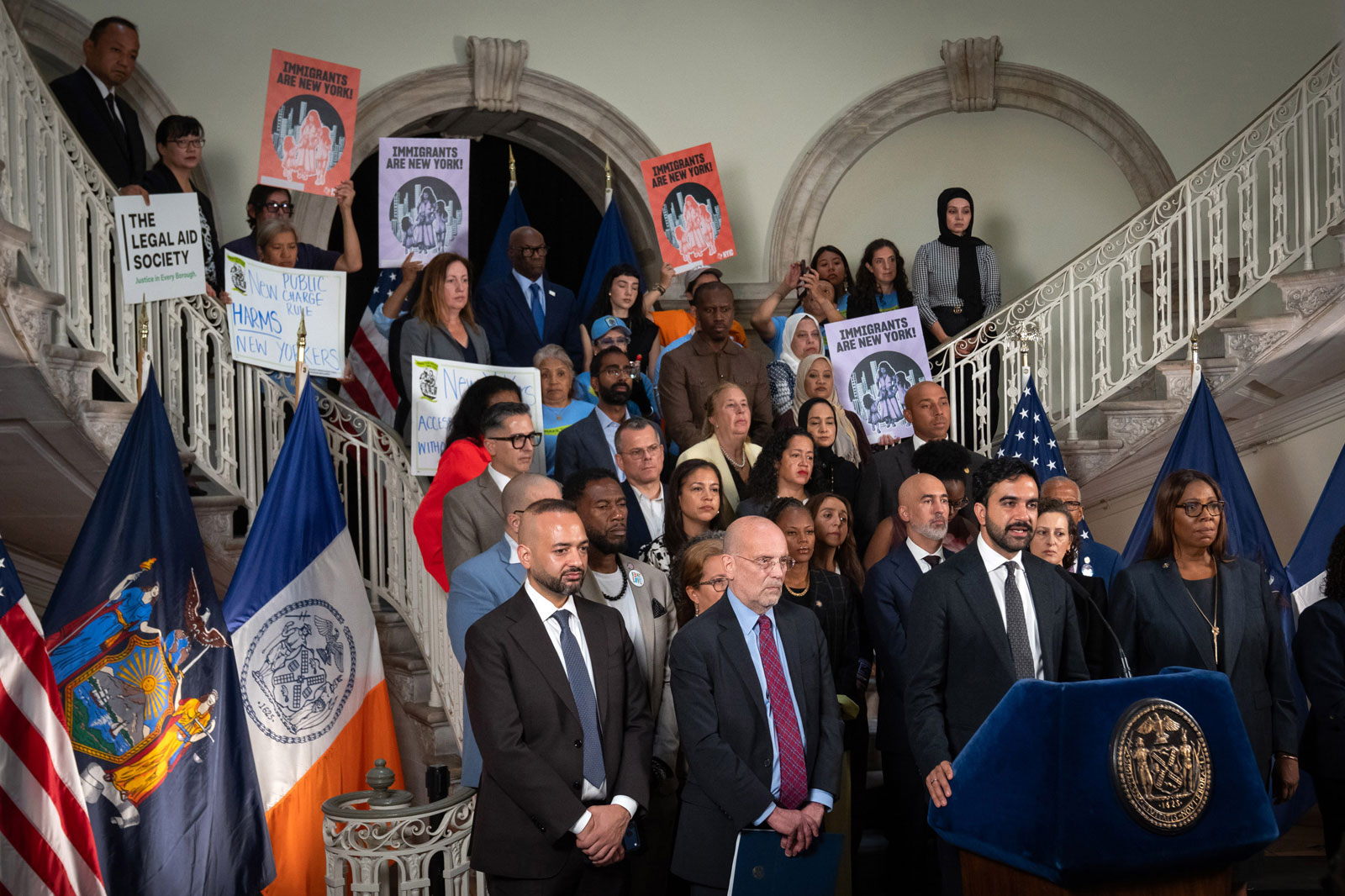 Mayor Mamdani, stands on staircase, with the podium in front, speaking to a crowd, while a group of people gather around hi,