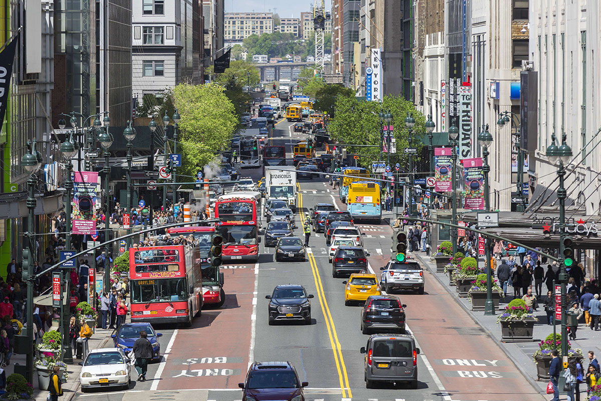Street in Manhattan with two lanes of traffic in either direction. Buses travel along the red bus lanes, while passenger vehicles travel in the center lanes