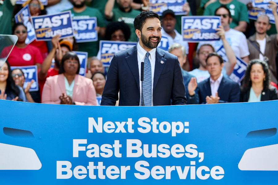 Mayor Zohran Mamdani stands at a podium in front of a crowd of people with a banner reading Next Stop: Fast Buses, Better Service in front of him during an outdoor press event.