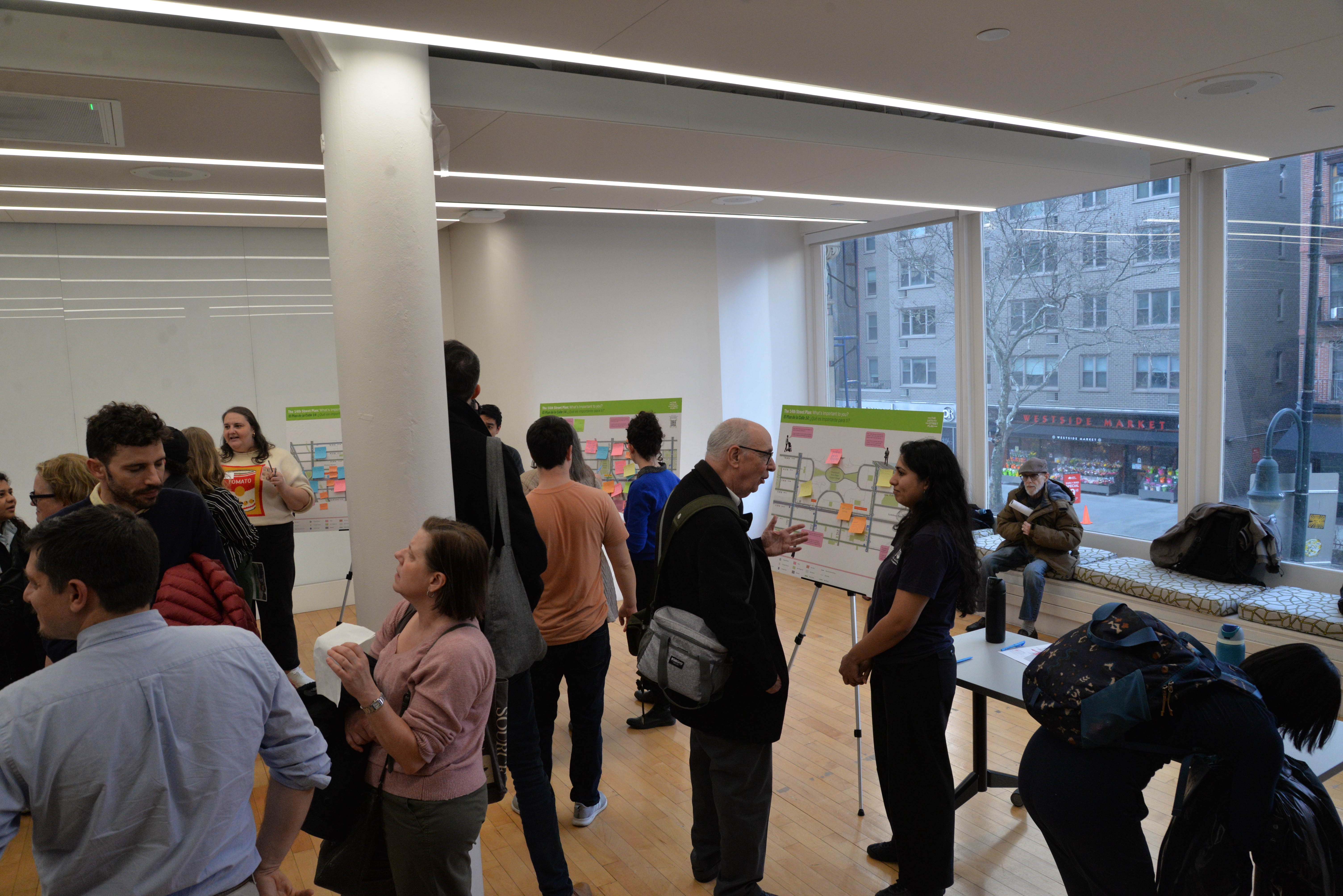 Multiple people speak with N Y C D O T representatives and add feedback via sticky notes on large boards at a public workshop.