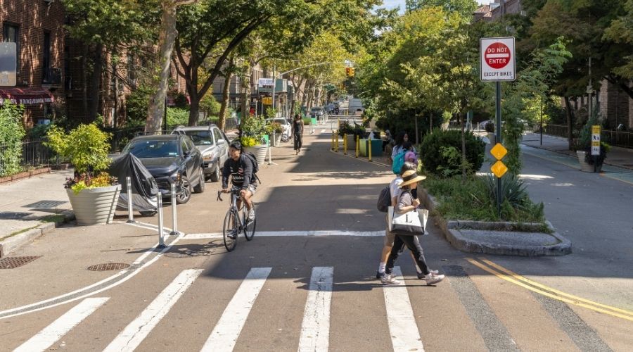 On a sunny day, people across a crosswalk in front of the pedestrianized 34th Avenue in Queens. The roadway is closed to vehicles and open for pedestrians and cyclists.