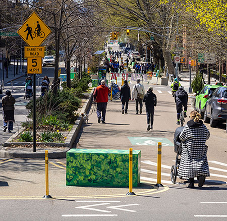 On a sunny spring day pedestrians of all ages stroll along a street closed to vehicles and open for walking and biking. A concrete block and yellow flexible posts create an extended median to slow down drivers when the roadway is normally open and shared by vehicles, cyclists, and pedestrians.