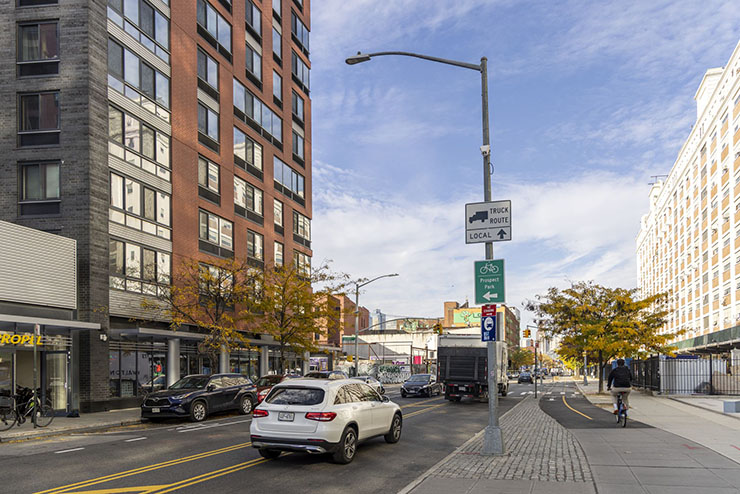 Cars and trucks travel along a roadway, with a raised bike lane and pedestrian area.  A small gray box is attached to a street light pole installed between the roadway and bike lane.