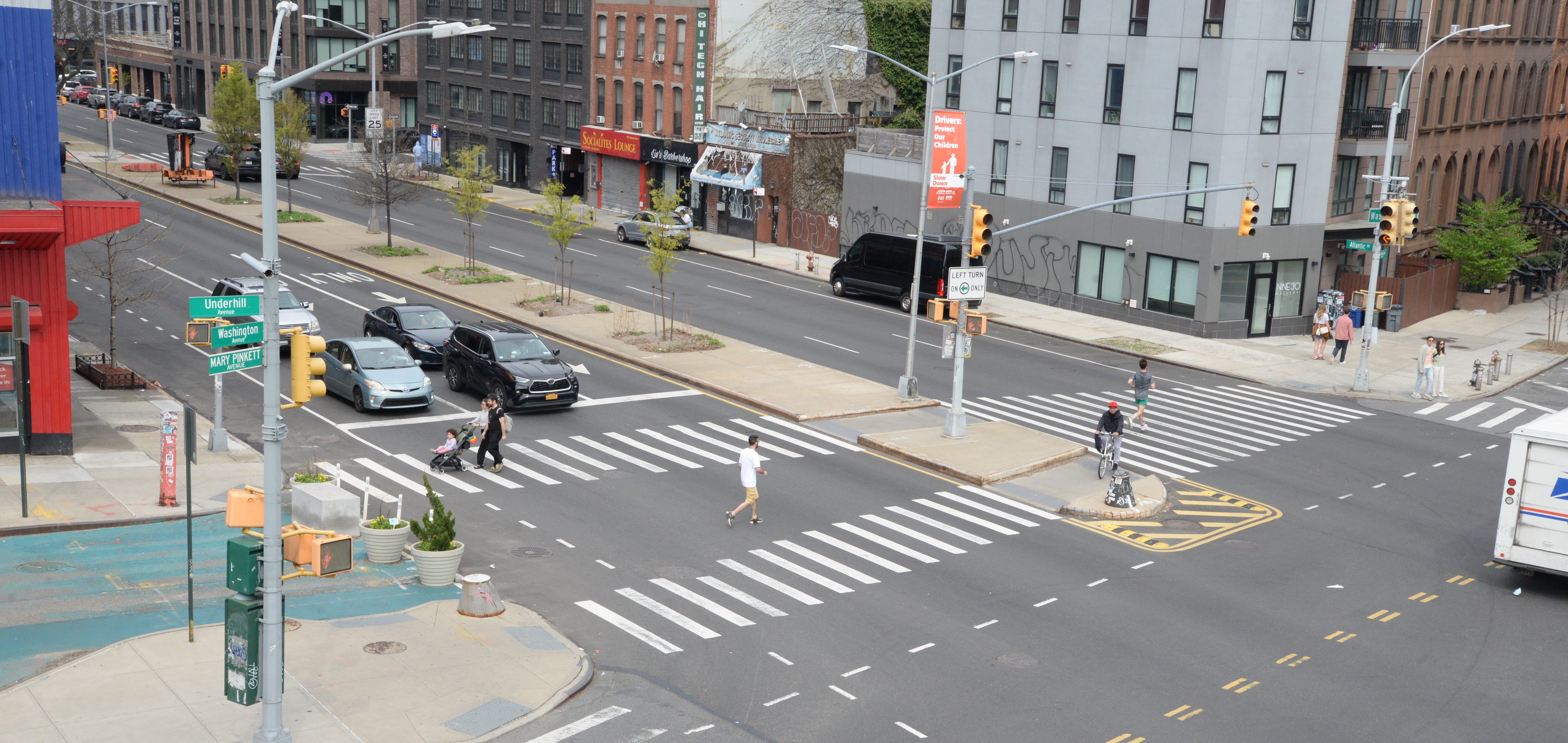  People walk, push strollers, and ride a bike in a wide crosswalk while drivers wait for pedestrians to cross Atlantic Avenue at Underhill Avenue in Brooklyn.