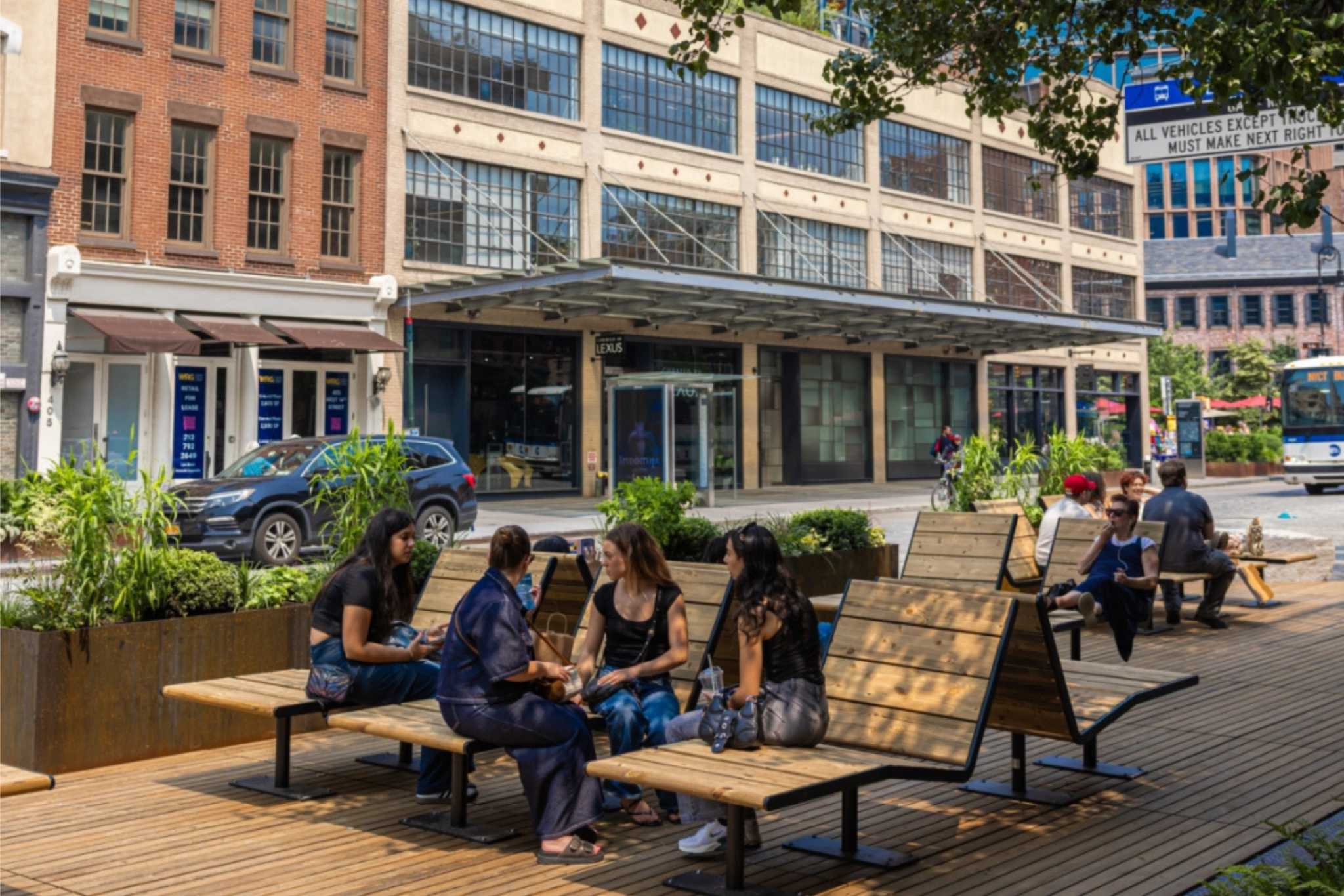People sit and relax on unique wooden lounge chairs installed on a custom wood plank platform on a pedestrianized section of 14th Street in Manhattan. An MTA bus travels along the vehicular section of the roadway.