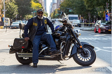 A man wearing motorcycle gear, leans on a motorcycle parked on a street closed to traffic.