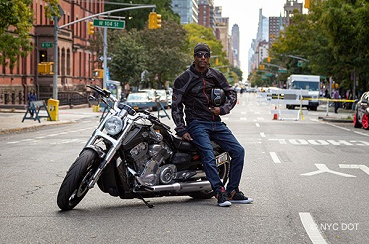 A man wearing motorcycle gear, holding a helmet, leans on a motorcycle parked on a street closed to traffic.