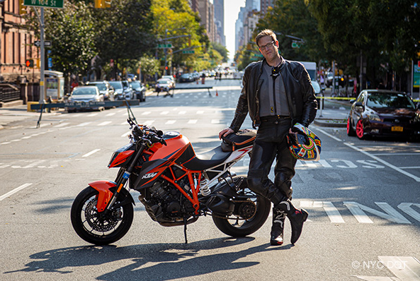 A man wearing motorcycle gear, holding a helmet, stands beside a motorcycle parked on a street closed to traffic.