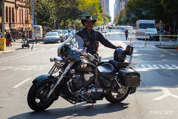 A man wearing motorcycle gear, holding a helmet, stands beside a motorcycle parked on a street closed to traffic.