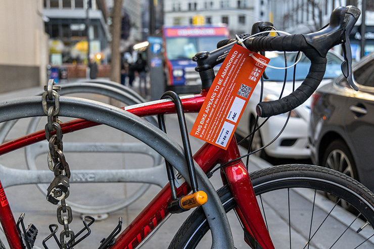 A red bike attached to a bike rack has an orange tag on the handlebars that says the bike appears to be abandoned