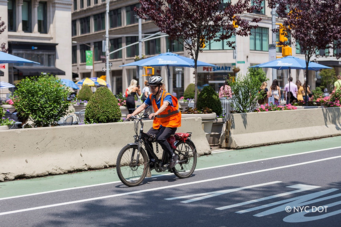 A delivery person riding an e-bike wears a helmet and orange safety vest while riding on a green bike lane.