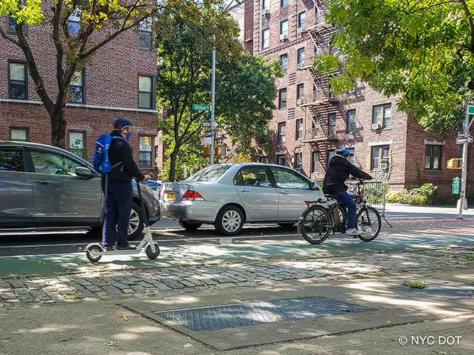 A person riding an e-scooter wears a helmet and backpack and travels along a green bike lane. Ahead of him is a cyclist riding an e-bike, wearing a helmet, riding along the green bike lane.