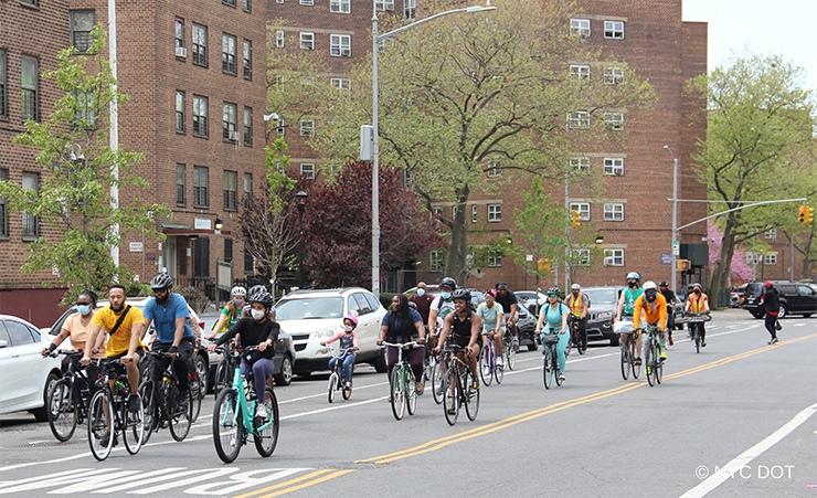 A large group of cyclists wearing helmets and face masks rides in a street in Brownsville, Brooklyn