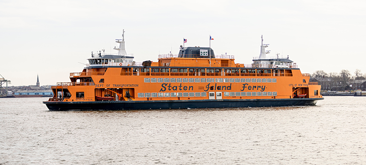 An orange ferry with blue script reading "Staten Island Ferry" travels in the New York Harbor on an overcast day.