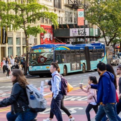Multiple pedestrians and cyclist walk across Manhattan’s 14th Street. In the background, a bus drops off and picks up passengers at a bus stop.
