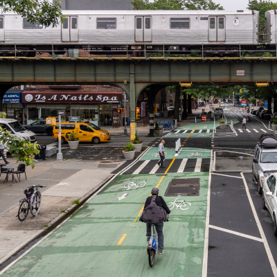 A cyclist bikes along a two-way bike lane painted green and at the intersection pedestrians walk in a crosswalk. The roadway and bike lane continues under an elevated subway train in Queens.
