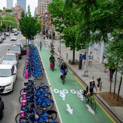 Multiple cyclists ride on a green bike lane, separated from vehicular traffic by a parking lane and a Citi Bike station.