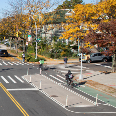 Multiple cyclists ride along a redesigned street in Queens featuring parking-protected green bike lanes along the curb.