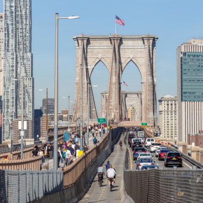 Multiple cyclists, pedestrians, and vehicles travel across the Brooklyn Bridge.