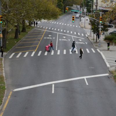 Pedestrians walk in a crosswalk across a three-lane wide roadway in Brooklyn.