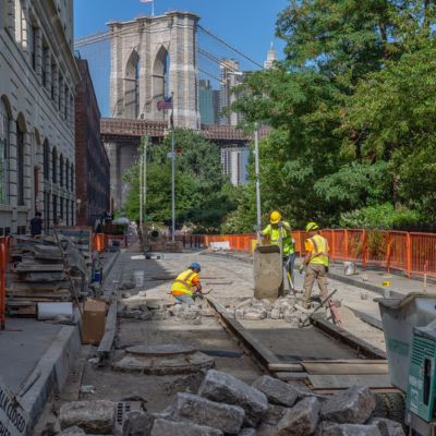 Workers remove and replace cobblestone in a Dumbo Street with the Brooklyn Bridge in the background.