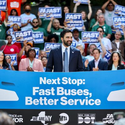 Mayor Mamdani makes an announcement before a large crowd. A banner in front of him reads Next Stop: Fast Buses, Better Service.