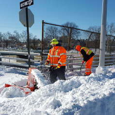 Two snow laborers wearing brightly colored clothing remove snow from a sidewalk.