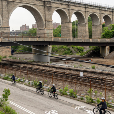 People ride bicycles along a paved path in the Bronx alongside railroad tracks with a bridge in the background.